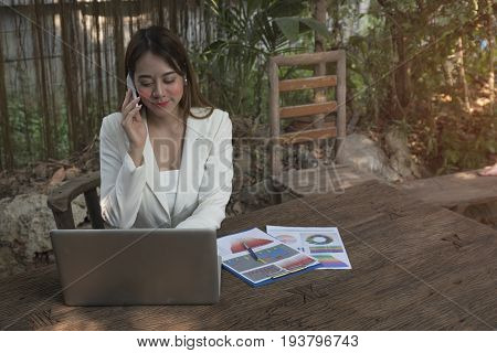 Asian smile beautiful girl sitting outdoor office using smart phone and laptop in nature. Work space with work sheet of business business and lifestyle conceptcopy space