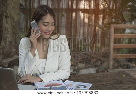Asian smile beautiful girl sitting outdoor office using smart phone and laptop in nature. Work space with work sheet of business business and lifestyle conceptcopy space
