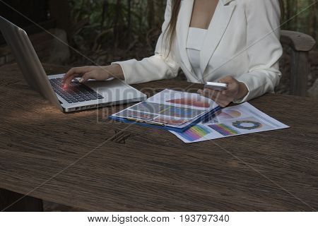 Asian smile beautiful girl sitting outdoor office using smart phone and laptop in nature. Work space with work sheet of business business and lifestyle conceptcopy space
