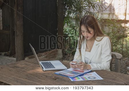 Asian smile beautiful girl sitting outdoor office using smart phone and laptop in nature. Work space with work sheet of business business and lifestyle conceptcopy space
