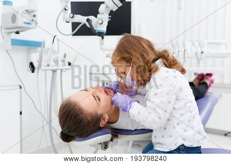 Adorable little girl sitting and making an examination of woman doctor lying on dentist chair in hospital. Dentist and child in cabinet. Dentist office. Little girl in the dentists office