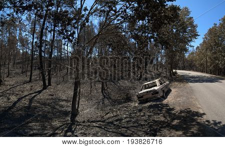 Road through the burnt forest and a car also completely burnt bye the side of the road. Pedrogao Grande Portugal.