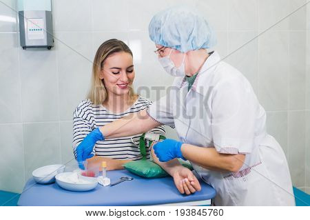 Preparation for blood test with pretty young blond woman by female doctor in white coat medical uniform on the table in white bright room. Nurse pierces the patient's arm vein with needle blank tube