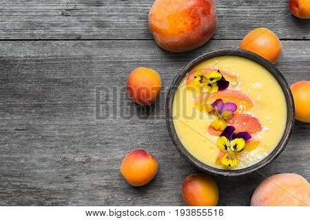 bowl of homemade yellow smoothie with fresh mango peaches and apricots topped with coconut and flowers over rustic wooden background. top view. healthy breakfast