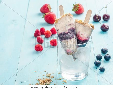 Homemade popsicle and cherry, blueberry, strawberry, raspberry on blue wooden background. Healthy summer breakfast. Popsicles from yogurt and banana with berries, granola and chia seeds. Copy space.