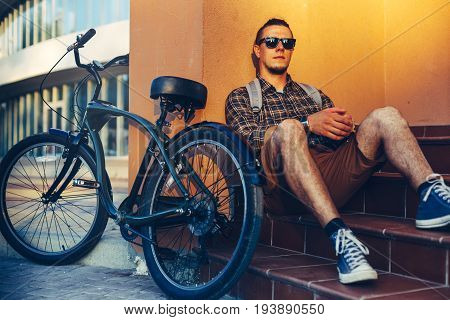 Handsome Guy Cyclist In Sunglasses With Backpack Sitting On The Steps Near The Bicycle Bike And Resting