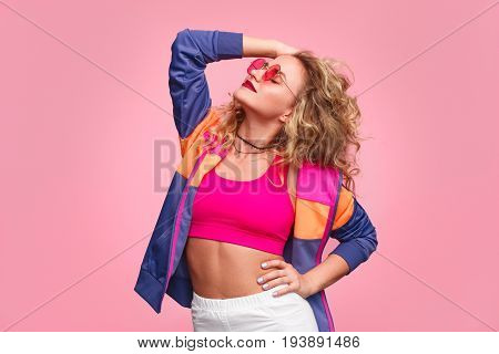 Blonde girl wearing pink glasses, pink top and blue jacket posing standing against pink background.