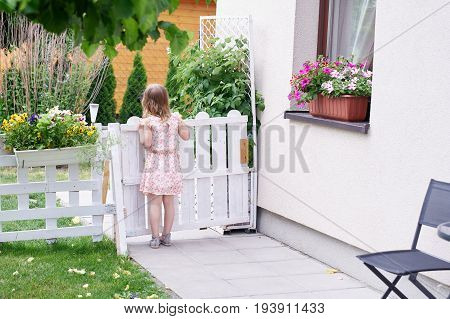 Little girl staying in a summer yard with her back to viewer and loooking with big interest outside the fence