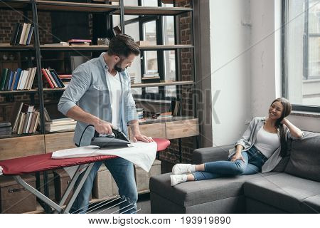 man ironing clothes while his girlfriend resting on sofa at home