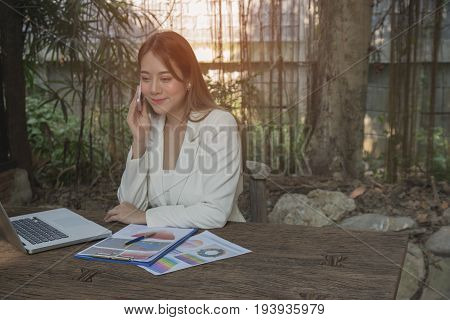 Asian smile beautiful girl sitting outdoor office using smart phone and laptop in nature. Work space with work sheet of business business and lifestyle conceptcopy space