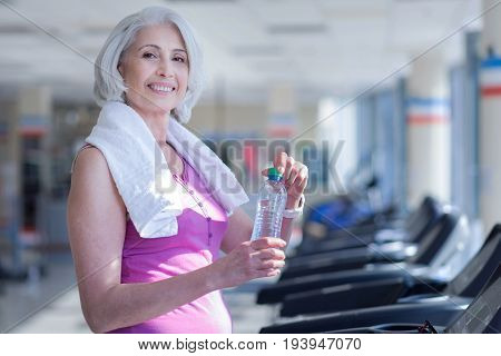 All you need. Smiling pretty senior woman covered with towel holding water bottle on race track at gym.