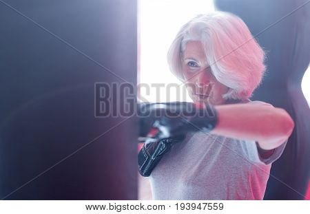 Strong hook. Close-up portrait of attractive strong aged lady standing and punching boxing bag at fitness studio.