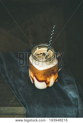 Iced caramel macciato coffee with milk in glass jar with straw over dark rustic wooden table background, selective focus, copy space, vertical composition. Cold sweet refreshing summer drink concept