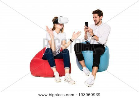 Smiling friends experiencing virtual reality glasses seated on beanbags isolated on white background. Experiencing virtual gaming adventure. VR