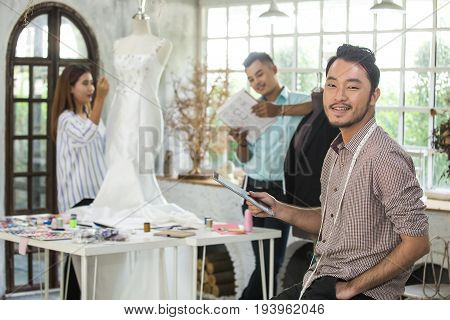 Young Asian Man Desiner Using Tablet For Design Woman Dress With Team.