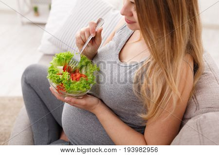 Young pregnant woman healthy eating, has green salad. Unrecognizable expectant blonde sitting on sofa and having fresh snack. Healthy nutrition and pregnancy concept