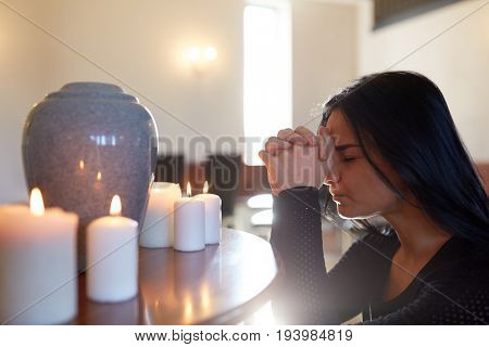 people and mourning concept - sad woman with cinerary urn and candles praying at funeral in church
