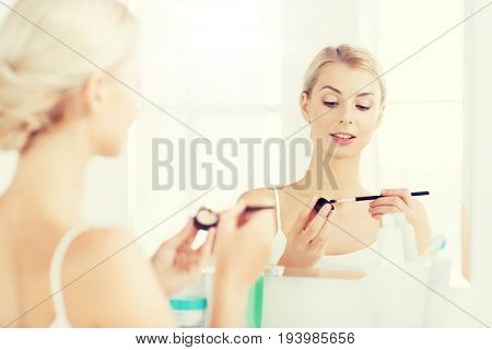 beauty, make up, cosmetics, morning and people concept - young woman applying eyeshade with makeup brush and looking to mirror at home bathroom