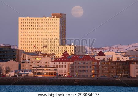 REYKJAVIK, ICELAND - MAY 03, 2015: Fosshotel building glowing reflecting sunset light, full Moon in the sky, snowy mountains in the background