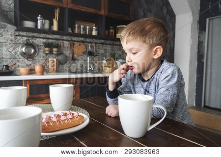 A Pensive Little Boy Sits At A Table With A Circley Tea And Cakes. Licking His Finger. Portrait.