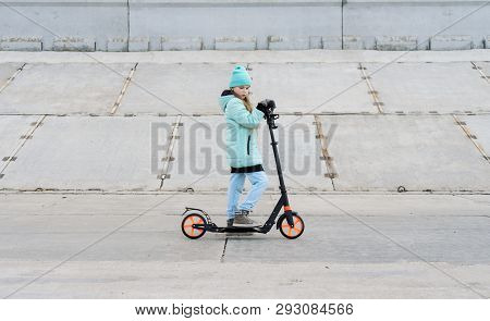 Activities: A School Girl With A Blue Jacket Riding A Scooter On A Gray Concrete Promenade.
