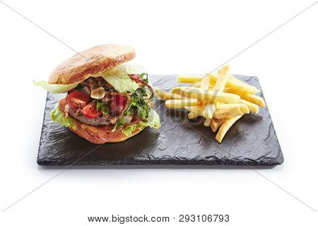 Fresh Beef Burger with Bacon, Fried Onions, Tomato Sauce, Pickled Cucumbers, Green Lettuce, Cheese and French Fries Garnish. Hamburger or Beefburger on Natural Black Stone Plate Isolated on White