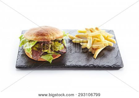 Classic Roast Beef Burger with Mustard, Fried Onions, Lettuce, Green Salsa Verde Sauce and French Fries Garnish. Beefburger with Medium Rare Steak on Natural Black Stone Plate Isolated on White