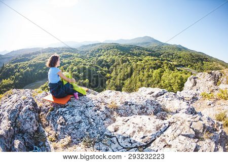 A Woman Is Sitting In A Sleeping Bag At Sunset. Camping On Top Of A Mountain. A Smiling Girl Rests A