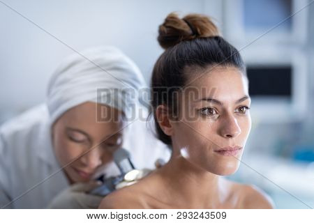 Front view of a mature mixed race female doctor checking a thoughtful Caucasian patient with dermatoscopy in clinic