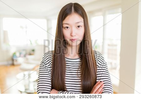 Beautiful Asian woman wearing stripes sweater over living room background Relaxed with serious expression on face. Simple and natural with crossed arms