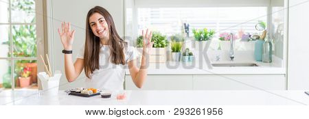 Wide angle picture of beautiful young woman eating asian sushi from delivery showing and pointing up with fingers number eight while smiling confident and happy.