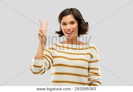 gesture, counting and people concept - happy smiling young woman in striped pullover showing peace hand sign or two fingers over grey background