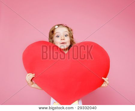 Lovely curly little girl holding large paper heart, over pink background