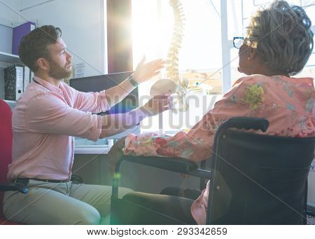 Side view of confident Caucasian male doctor illustrating his words by showing an anatomical model to senior woman in clinic