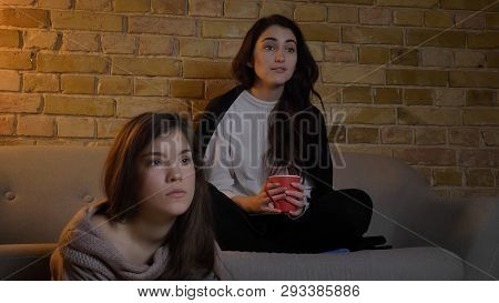 Closeup Portrait Of Two Young Female Friends Watching Tv While Resting In A Cozy Apartment Indoors