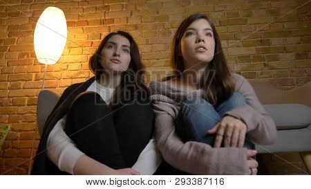 Closeup Bottom Up Portrait Of Two Young Pretty Women Watching A Tv Show In A Cozy Apartment Indoors