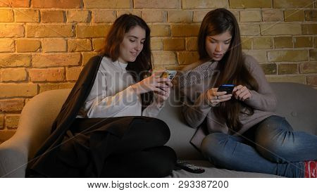Closeup Portrait Of Two Young Pretty Caucasian Girls Using The Phones While Resting On The Couch Ind