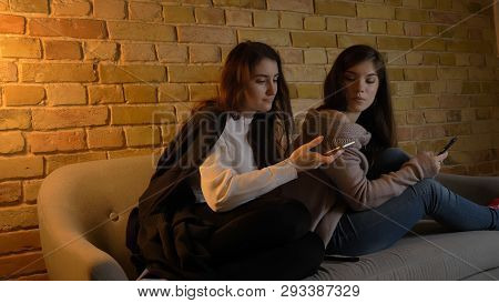 Closeup Portrait Of Two Young Pretty Caucasian Girls Using The Phones While Resting On The Couch Ind