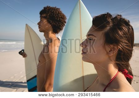 Side view of thoughtful young multi-ethnic couple standing on the beach with their surfboards and watching the waves at beatch