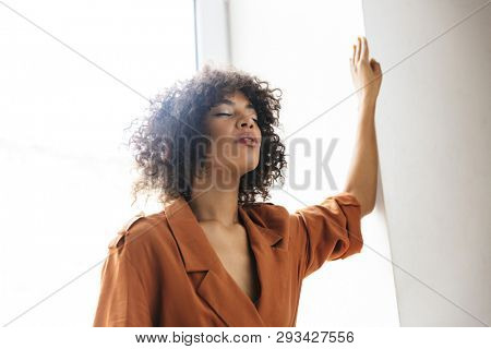 Image of Meditator pretty african woman posing with closed eyes near the wondow