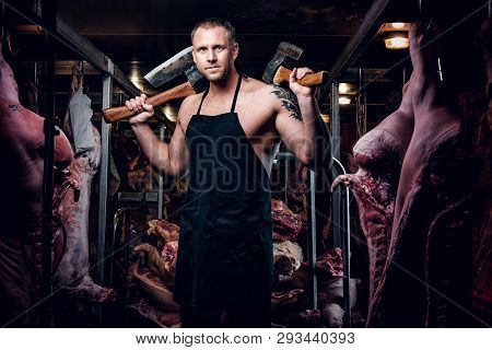 Strong Butcher With A Tattoo On His Hand Wearing Apron Posing With Two Axes In A Refrigerated Wareho
