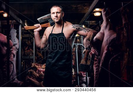 Strong Butcher With A Tattoo On His Hand Wearing Apron Posing With Two Axes In A Refrigerated Wareho