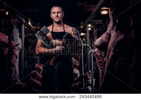 Strong Butcher With A Tattoo On His Hand Wearing Apron Posing With Two Axes In A Refrigerated Wareho