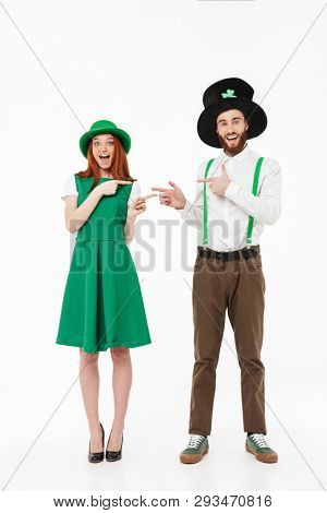 Full length of a happy young couple wearing costumes, celebrating St.Patrick 's Day isolated over white background, pointing fingers at each other