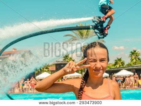 Smiling Teen Girl Enjoing Summer Vacation At The Pool With Fly Board Watershow On The Background. Gi