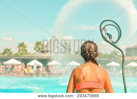 Teen Girl Watching Fly Board Watershow At The Pool. View From The Back. Summer Holidays, Fun Summert