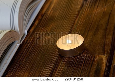 A Burning Candle Stands On Wooden Planks Next To An Open White Book Of Both Religion And Faith. Clos