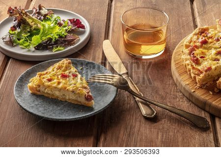 A French Dinner. A Slice Of A Quiche Lorraine With Mesclun Leaves And White Wine On A Dark Rustic Wo