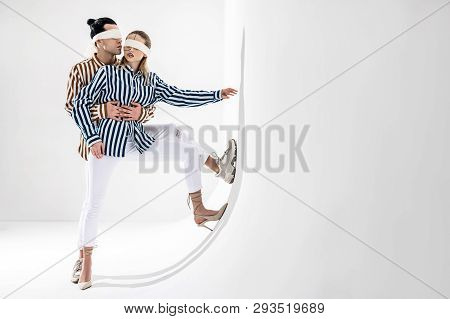 Couple Wearing White Trousers Posing Near White Wall In Blindfolds
