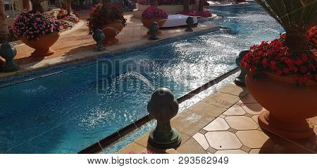 Fountain Jets Into The Water Canal Of Blue Outdoor Swimming Pool Surrounded By Exotic Flowers In Vas
